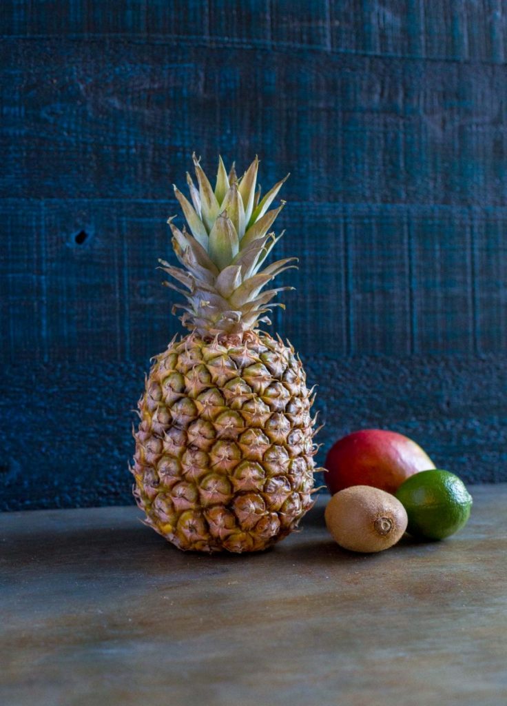 A tall pineapple next to a mango, lime and kiwi.