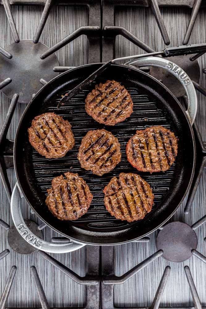 Six sliders with grill marks in a round Staub grill pan on a gas stovetop.