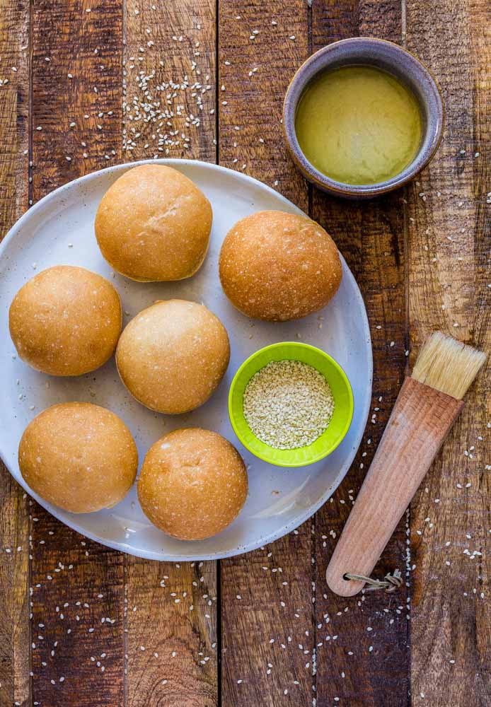 Six slider buns with a dish of sesame seeds, a pastry brush and melted butter.