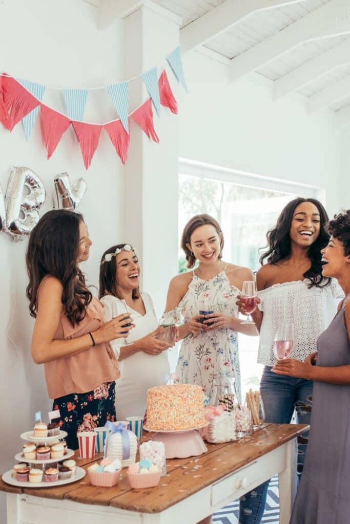 Five women at a baby shower socializing and playing games near a dessert table.