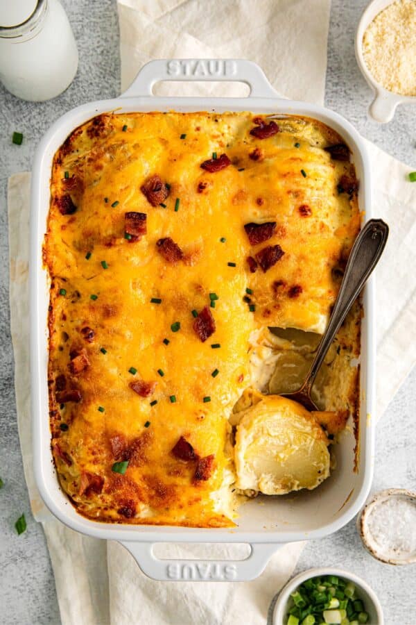Silver serving spoon digging into a baking dish of cheesy scalloped potatoes garnished with ham and bacon.