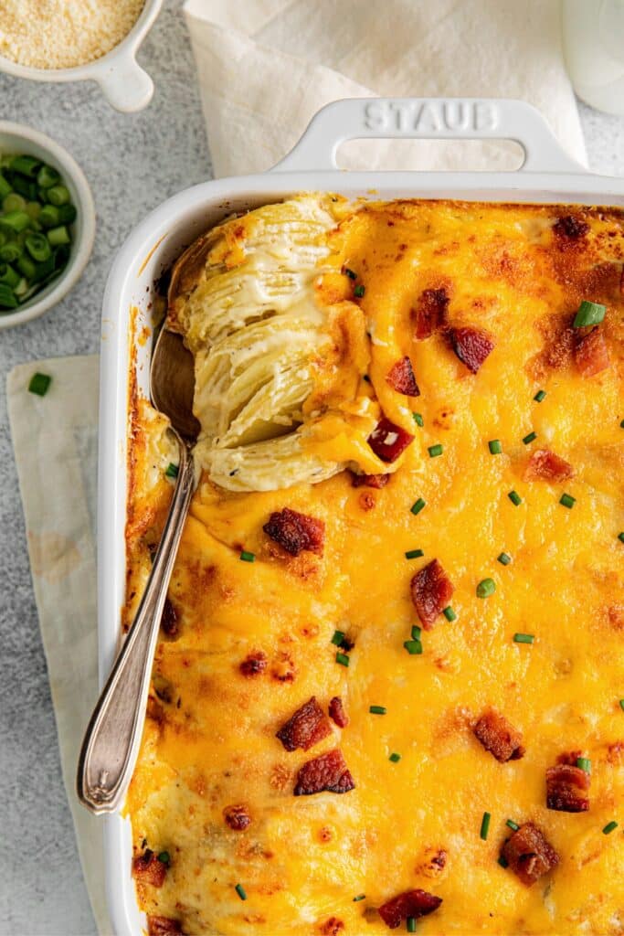 A serving spoon digging into a white baking dish of extra cheesy scalloped potatoes.