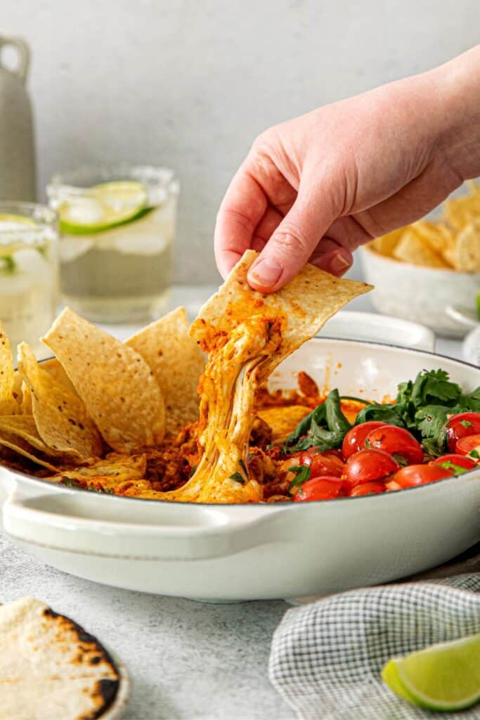 A hand holding a tortilla chip, scooping up a serving of queso fundido with chorizo.