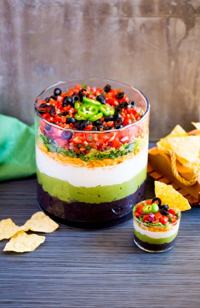 Mexican 7-layer dip in a large bowl and in a mini bowl with crispy tortilla chips.