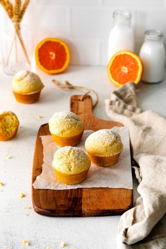Orange muffins sprinkled with powdered sugar next to fresh oranges and bottles of milk.