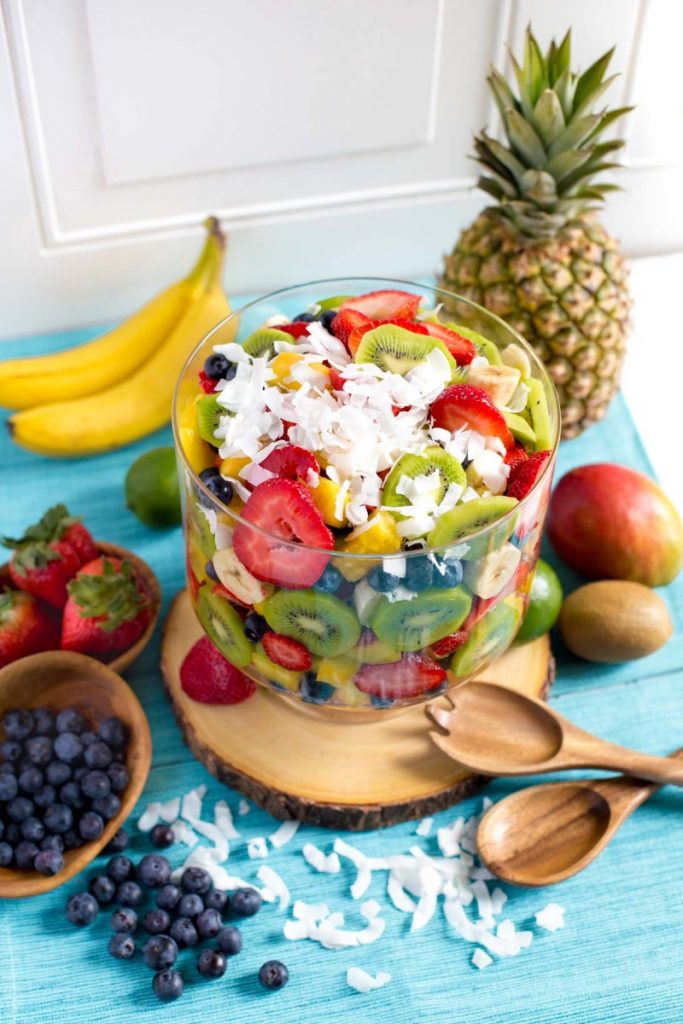 Tropical fruit salad in a glass trifle bowl with wooden salad servers.