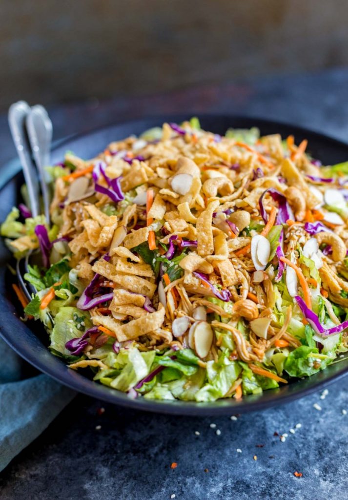 A close-up view of chicken salad in a black wok-shaped bowl with serving spoons.