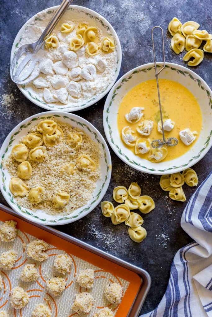 Three shallow bowls with flour, egg and breadcrumbs for prepping baked tortellini appetizers.