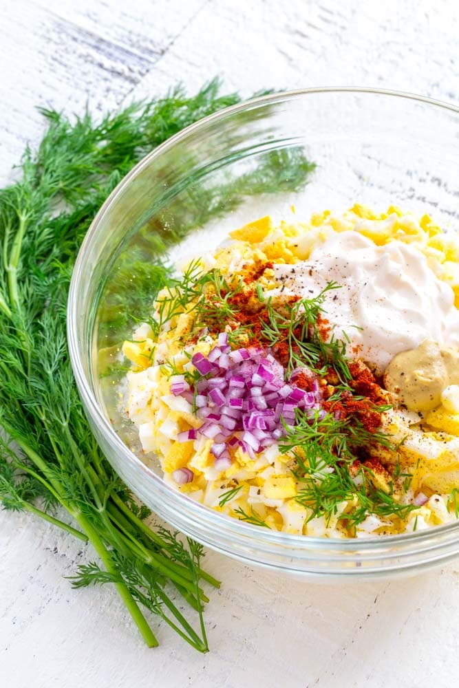 Egg salad ingredients in a clear glass mixing bowl next to sprigs of fresh dill.