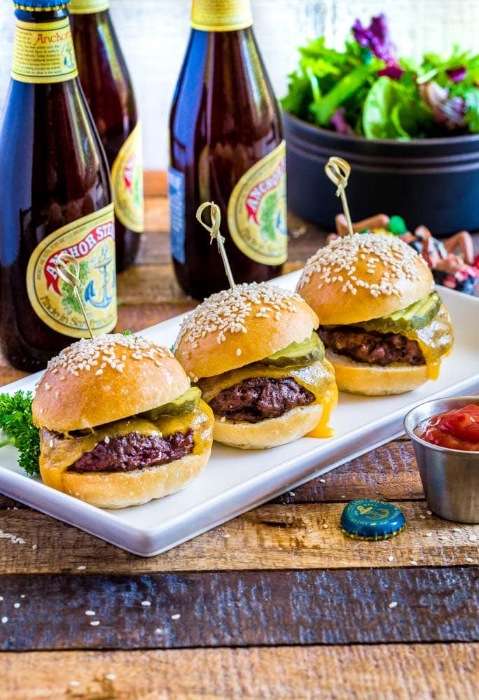 Three cheeseburger sliders on a white rectangular platter with craft beer in the background.