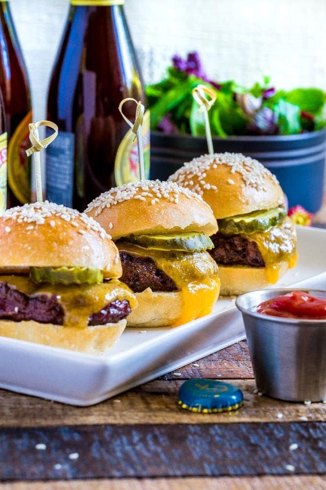 Three cheeseburger sliders on a thin rectangular platter with a pickle and bamboo pick.