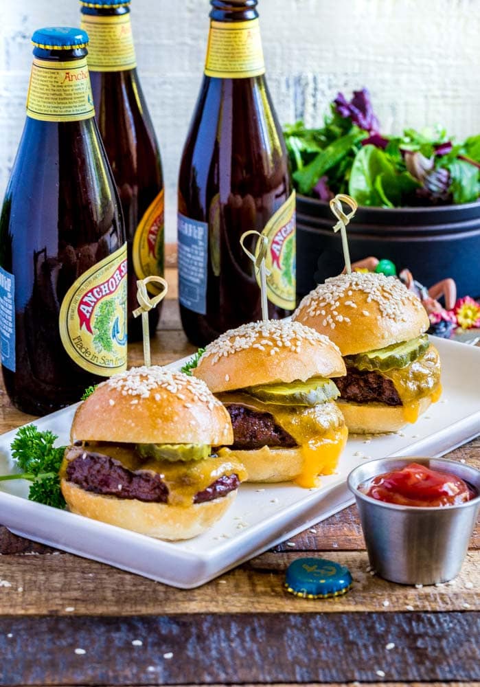 Three cheeseburger sliders on a thin rectangular platter with a pickle and bamboo pick.