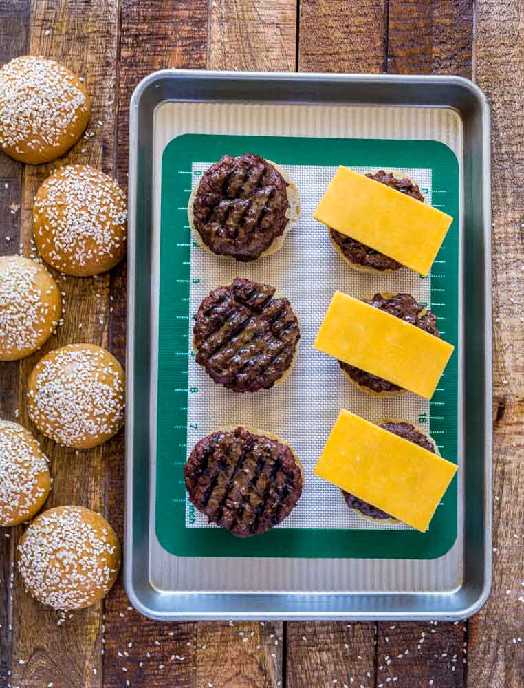 Six seeded buns next to baking tray of open-faced cheeseburger sliders.