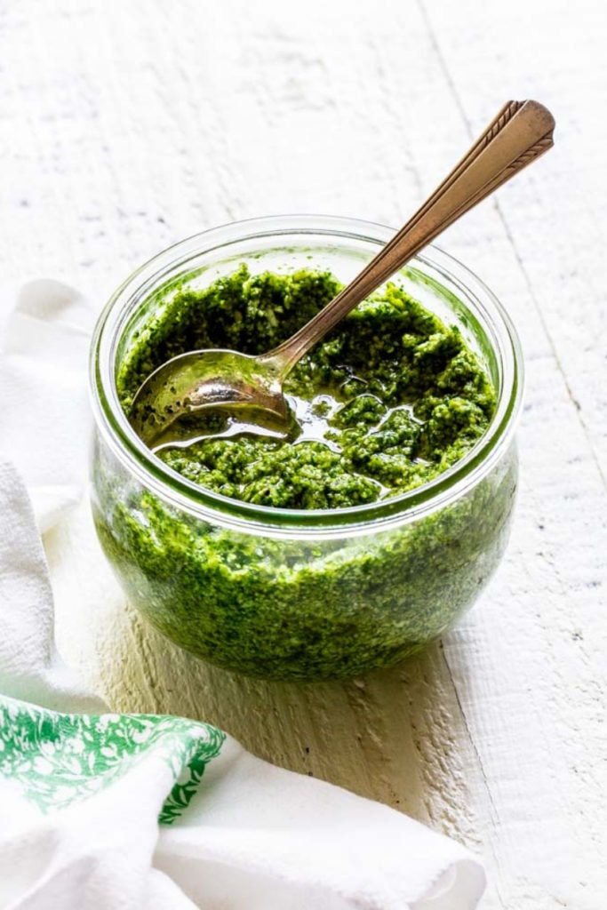 Basil pesto in a Weck glass bowl with a silver spoon inside on a white table.