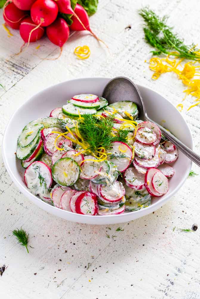 Cucumber salad in a white bowl with a silver serving spoon.