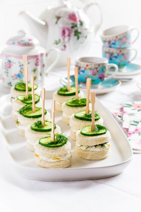 Cucumber tea sandwiches on a white serving tray surrounded by a tea pot and tea cups.