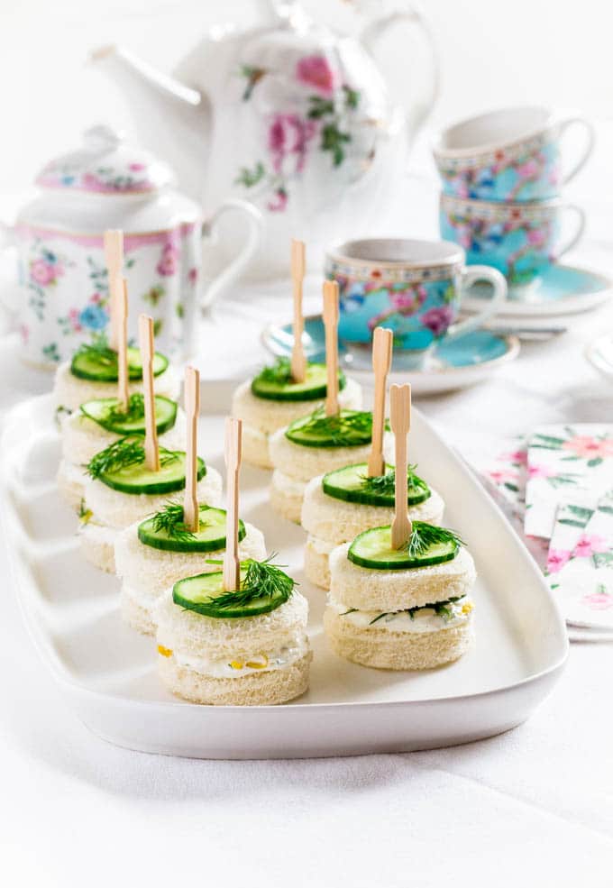Cucumber tea sandwiches on a white serving tray surrounded by a tea pot and tea cups.