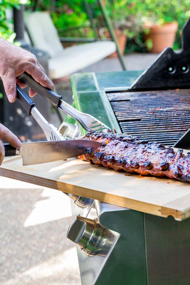 A rack of BBQ Ribs being sliced with a knife on a wooden cutting board.