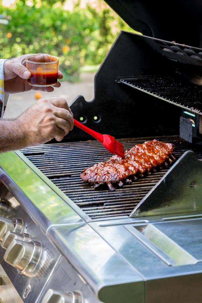 A man standing in front of an outdoor grill slathering BBQ sauce on baby back ribs.
