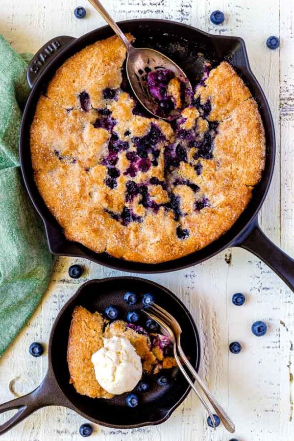 A Staub cast-iron skillet with blueberry cobbler and a large metal serving spoon.
