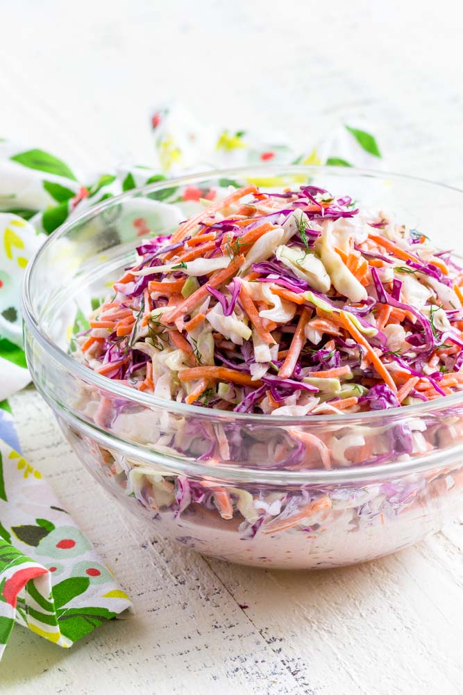 Coleslaw recipe presented in a clear serving bowl next to a flower-printed flour-sack towel.