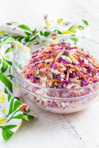 A clear bowl filled with colorful coleslaw next to a flour sack towel on a white farm table.