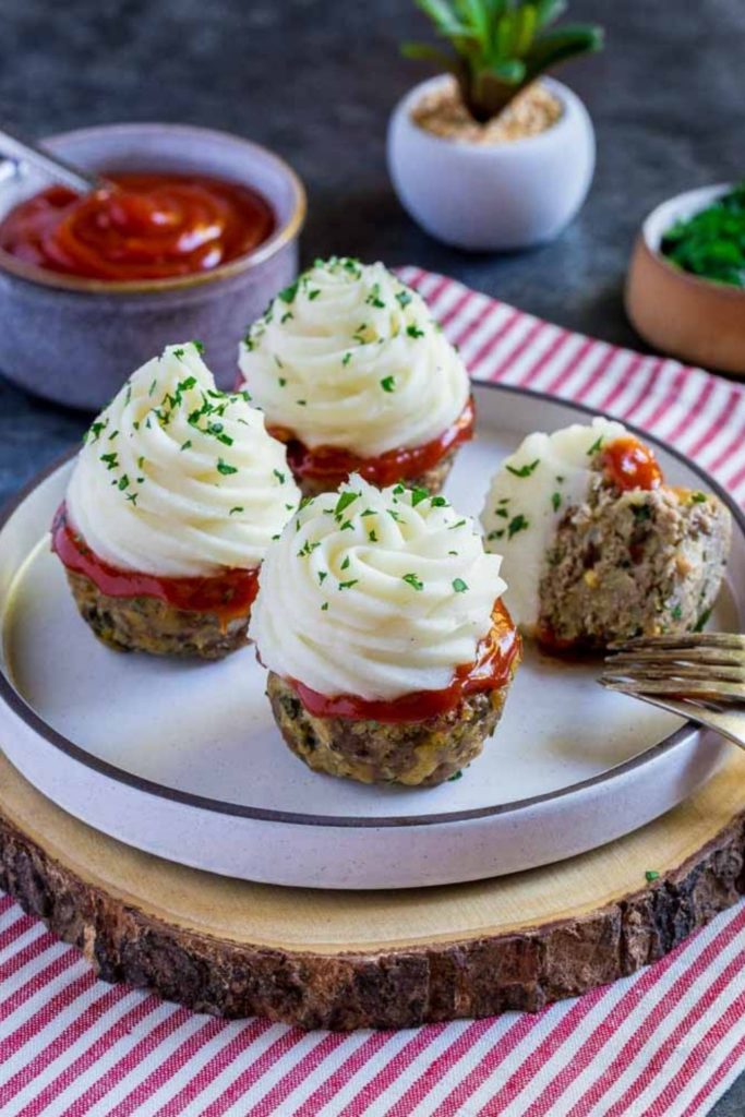 Four meatloaf cupcakes on a small round serving dish next to a dish of ketchup.