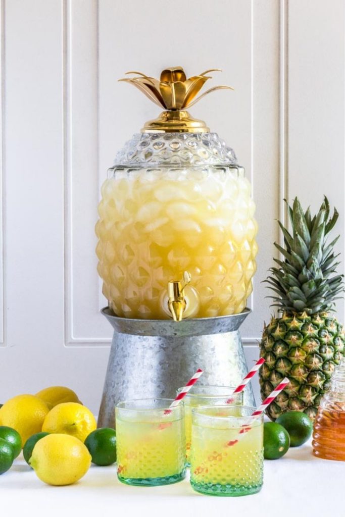 Rum punch recipe served in a glass pineapple beverage dispenser on a metal stand.
