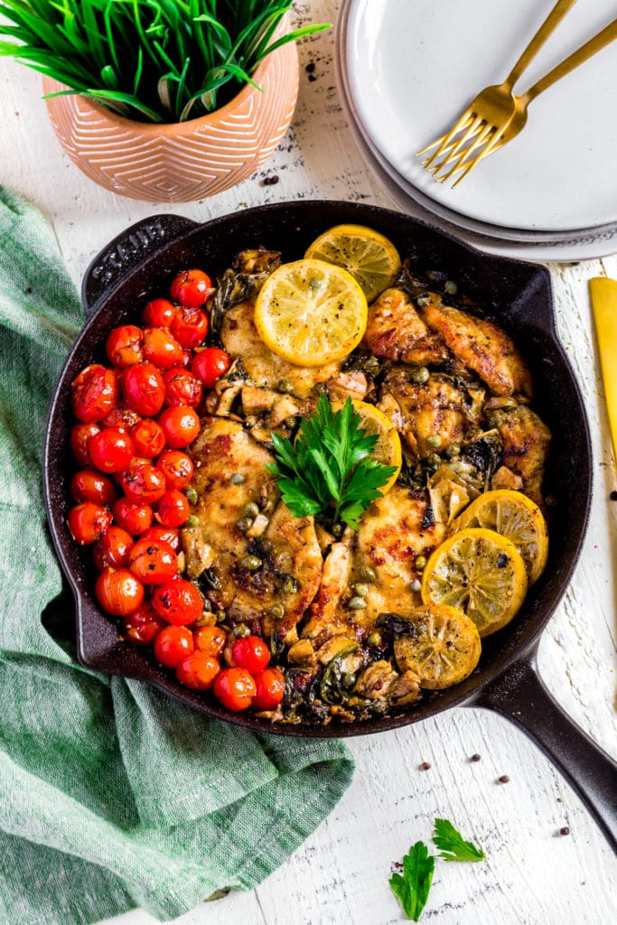 Chicken piccata with lemon and capers in a skillet next to dinner plates and forks.