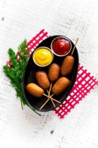 Homemade corn dogs served in an oblong metal restaurant tray with sides of mustard and ketchup.