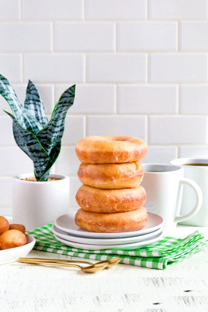 A stack of 4 glazed donuts next to 2 cups of coffee.