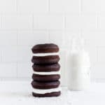 A stack of three whoopie pies next to 2 glass bottles of milk.