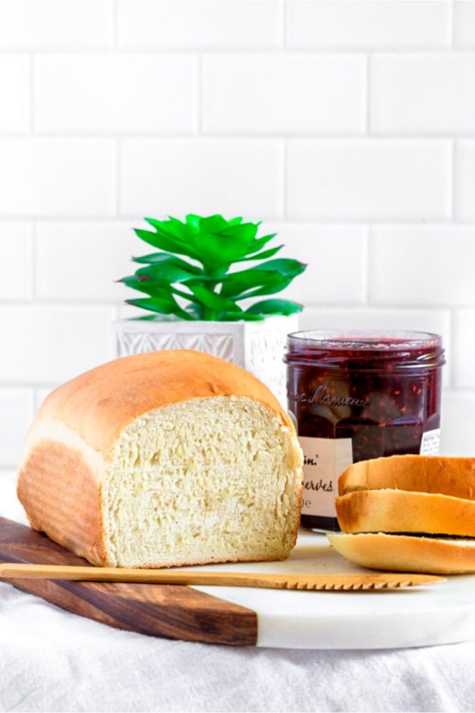 Fresh loaf of homemade white bread on a marble-and-teak board next to a jar of preserves.