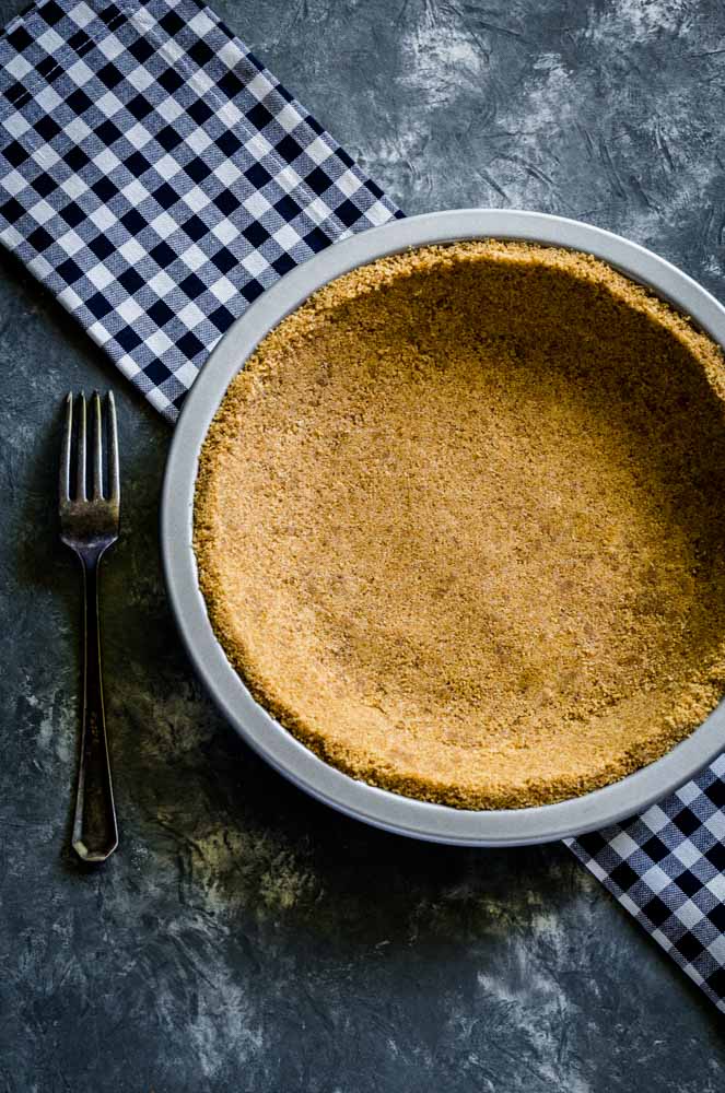 Graham cracker crust in a metal pie pan next to a tarnished metal fork.