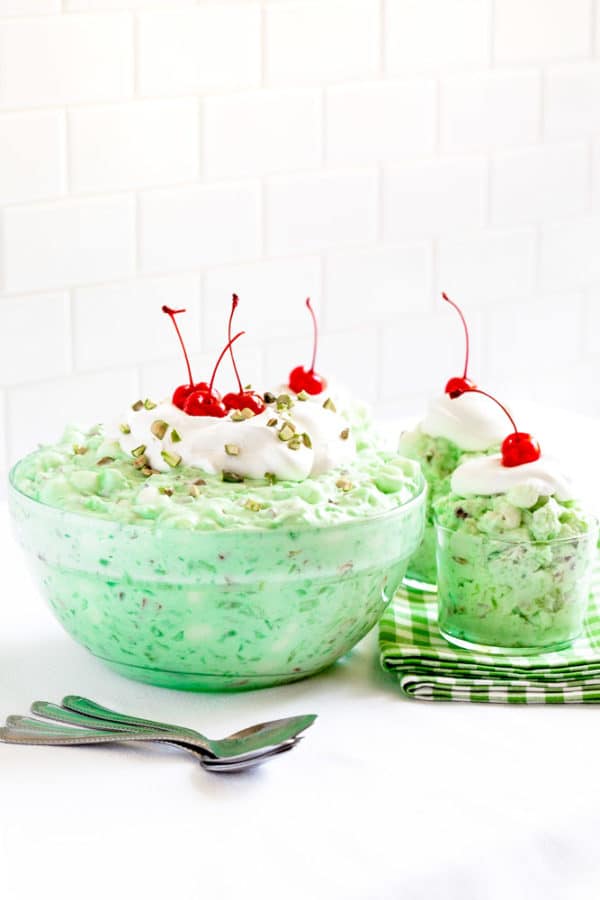 A large glass bowl of Watergate Salad with dessert cups garnished with whipped cream and a cherry.