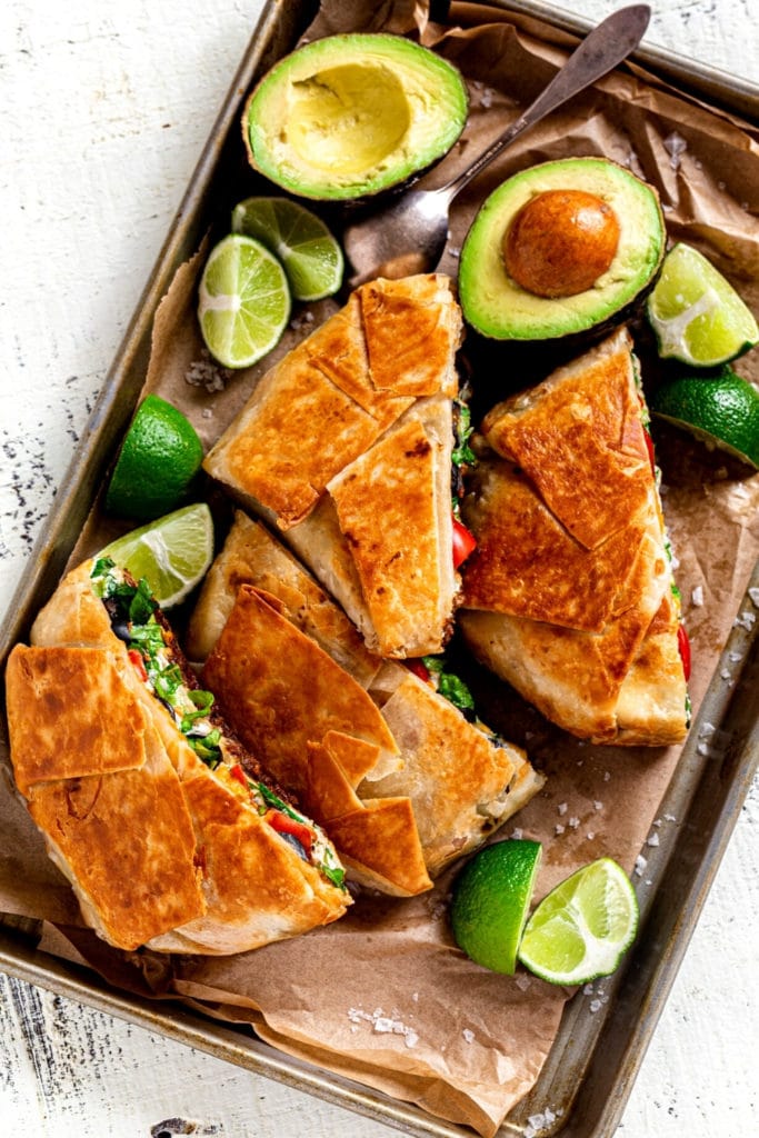 Two crunchwrap supreme entrees on a metal serving tray with avocados and limes.