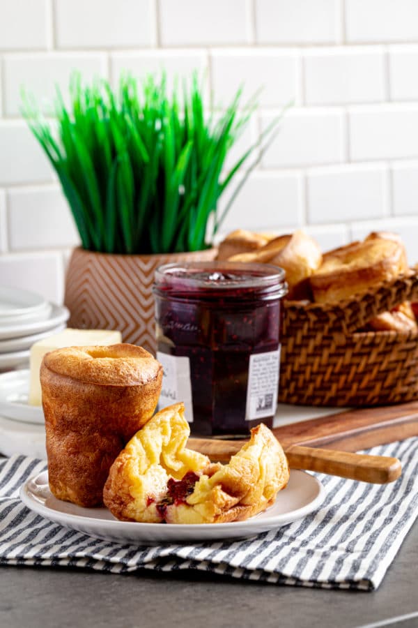 Two popovers on a plate next to a jar of jam an wicker basket with popovers.