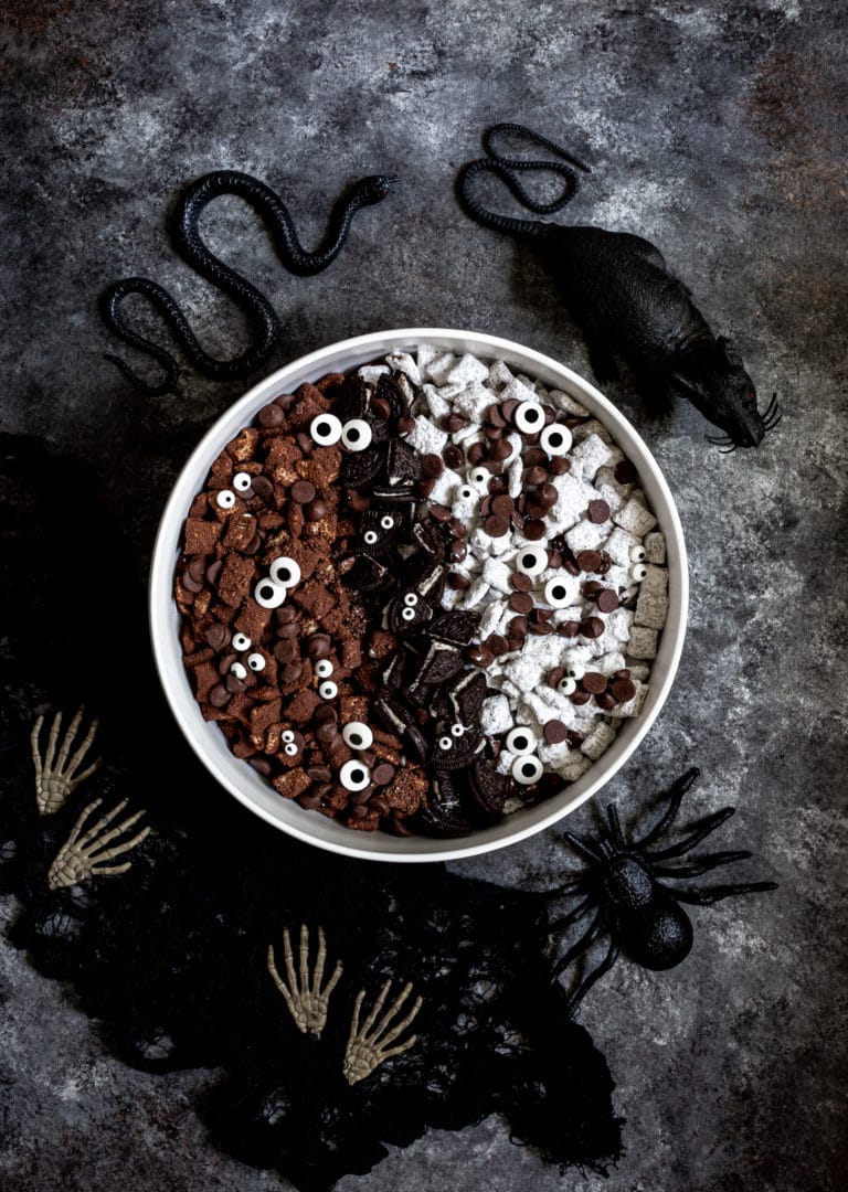 Halloween muddy buddies (chocolatey snack mix) served in a white bowl surrounded by spooky props.