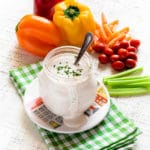 Homemade buttermilk ranch dressing in a mason jar surrounded by fresh veggies: bell peppers, carrots, baby tomatoes, celery.