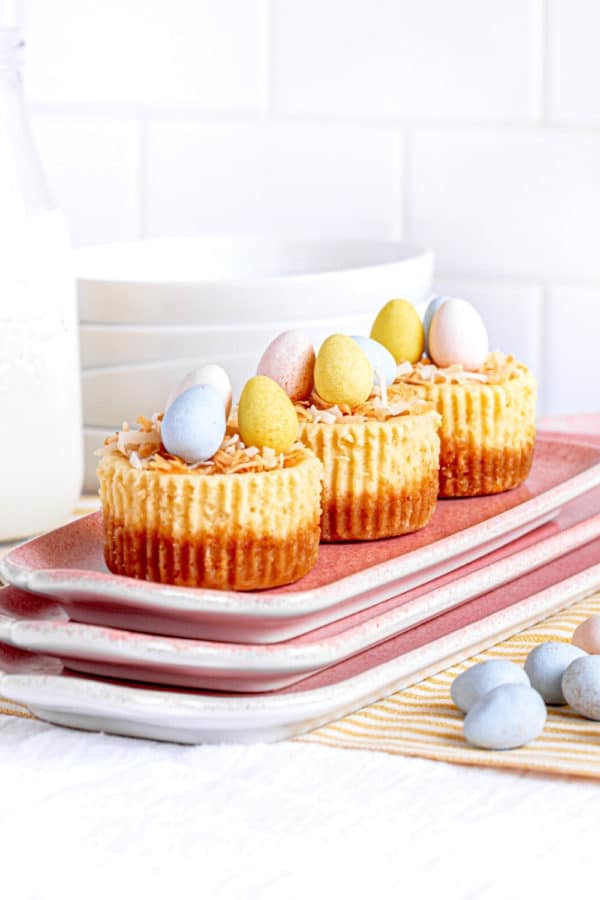 Mini Easter cheesecakes with a bird's nest of shredded coconut with candy eggs on a pink platter.