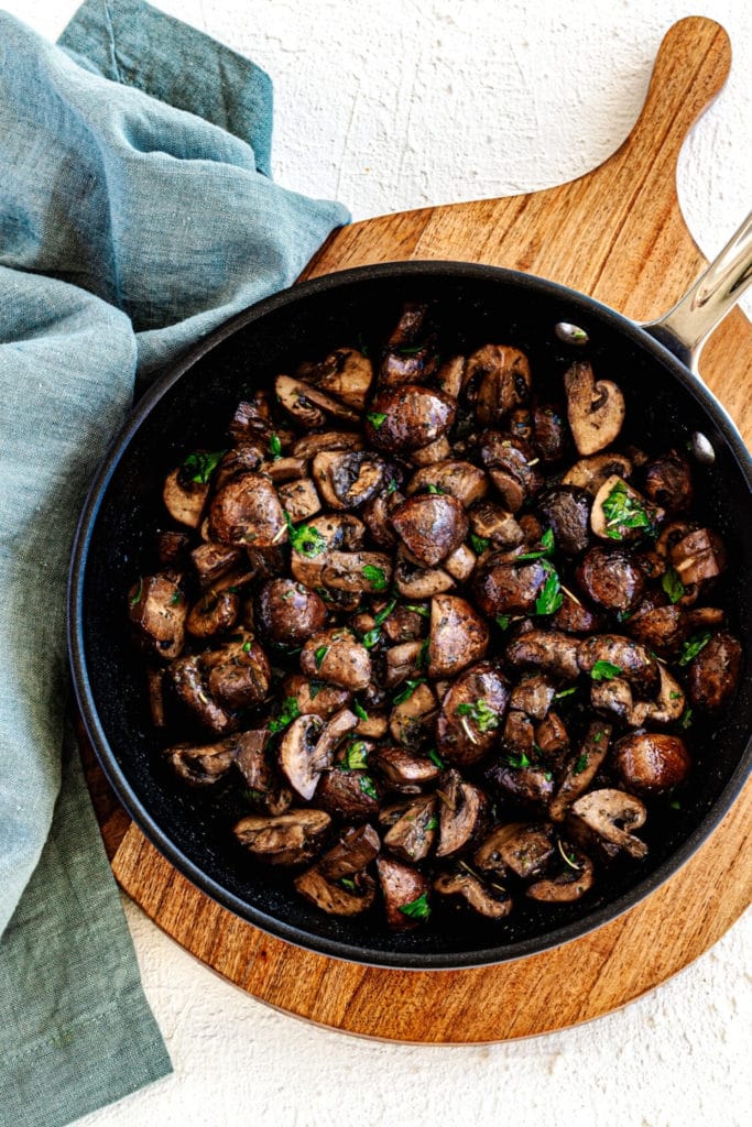 Heavy skillet with sauteed mushrooms garnished with chopped parsley on a wooden trivet.