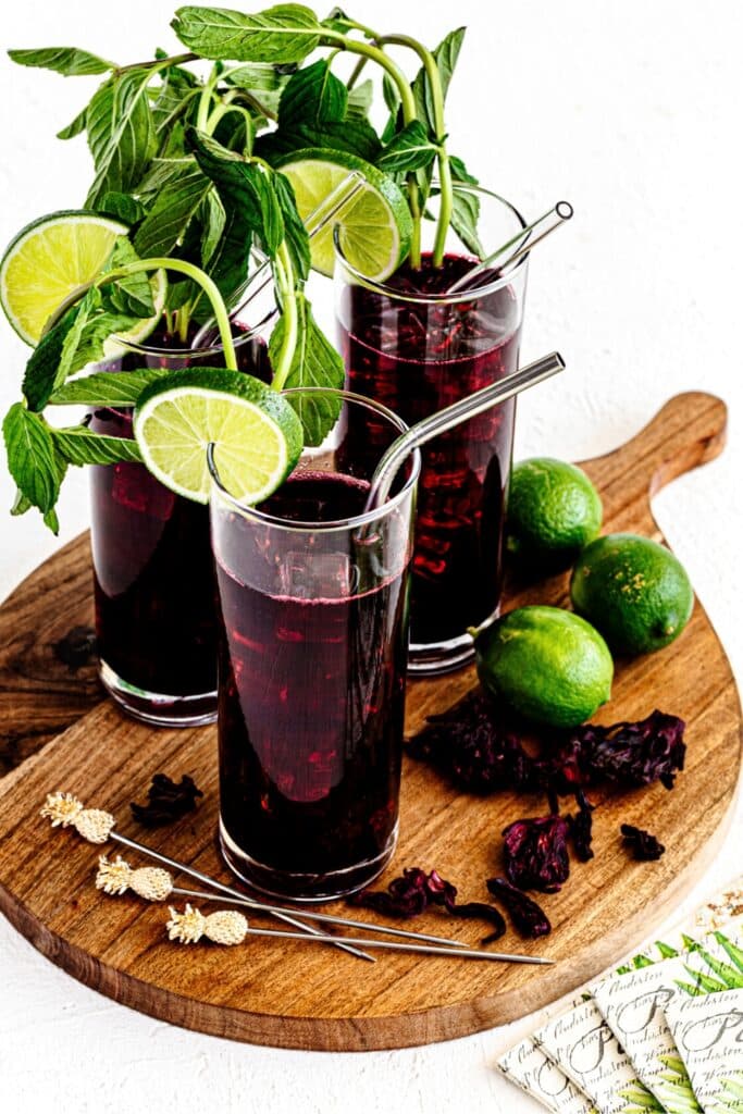 A wooden serving platter with 3 glasses of agua de Jamaica garnished with fresh mint and lime.