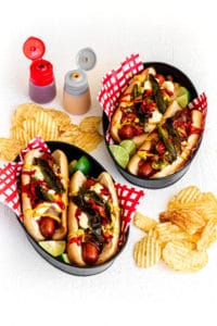 Two metal food baskets with Sonoran Hot Dogs (Mexican Hot Dogs) with condiment bottles and chips.