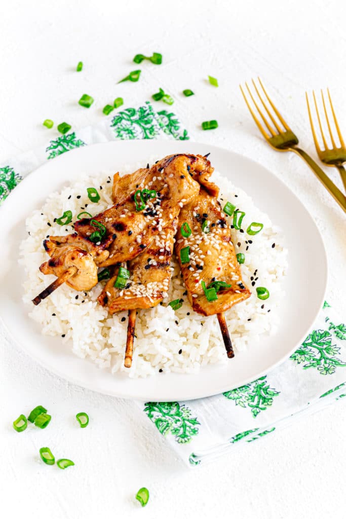 Teriyaki chicken on a stick over fluffy white rice sprinkled with sesame seeds and green onions.