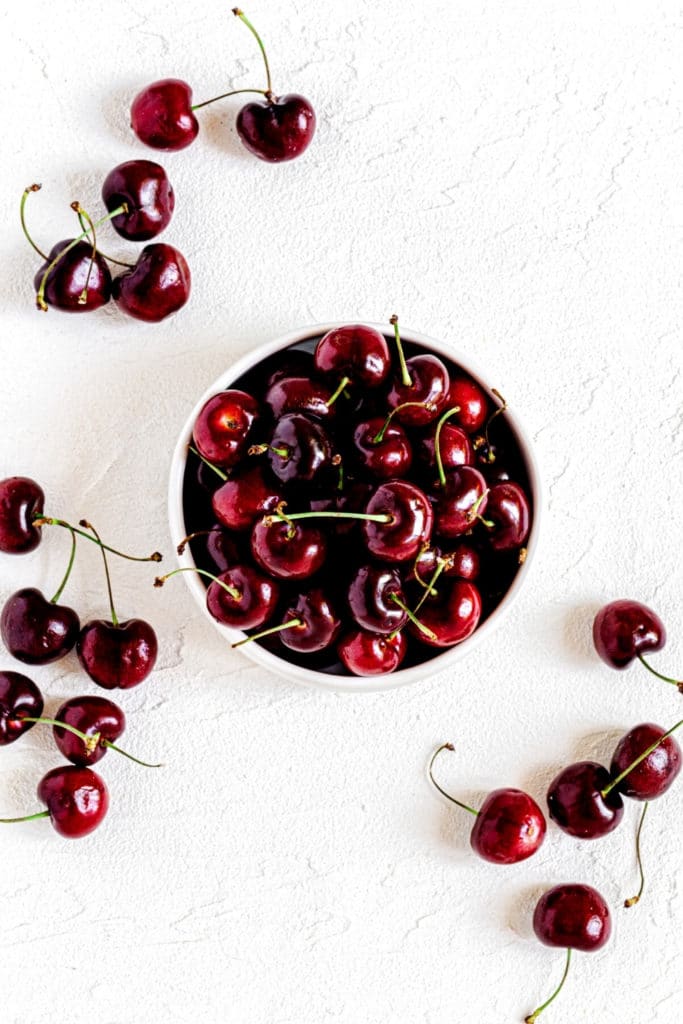 A bowl of fresh cherries with stems on a white surface.