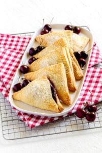 Six homemade cherry hand pies on a white platter next to a scattering of fresh cherries.
