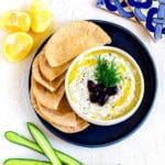 Tzitziki Sauce garnished with dill and Kalamata olives, served with pita bread on a blue platter.