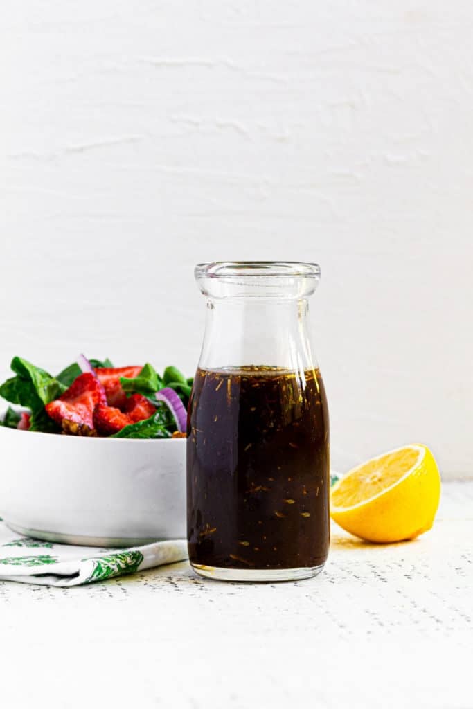 A salad dressing bottle with creamy balsamic dressing next to a bowl of strawberry salad.