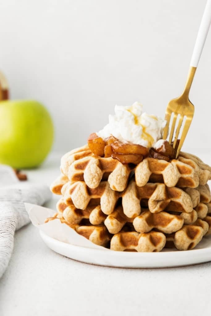 Stack of freshly-made apple cinnamon waffles on a white plate with gold fork.