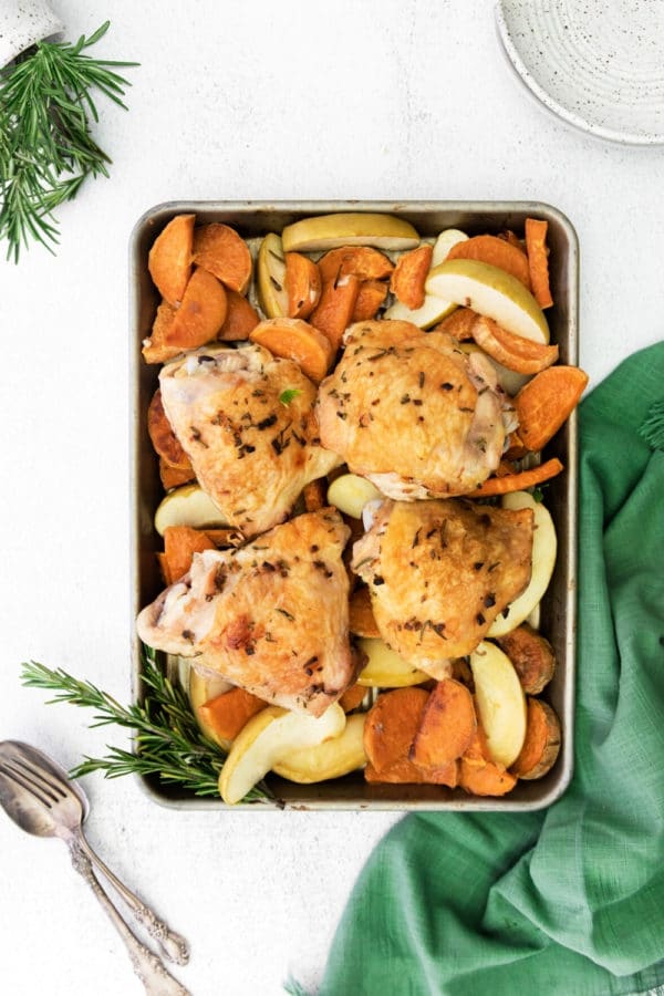 Chicken sweet potato bake sheet pan dinner with apples and rosemary on a tabletop with plates.